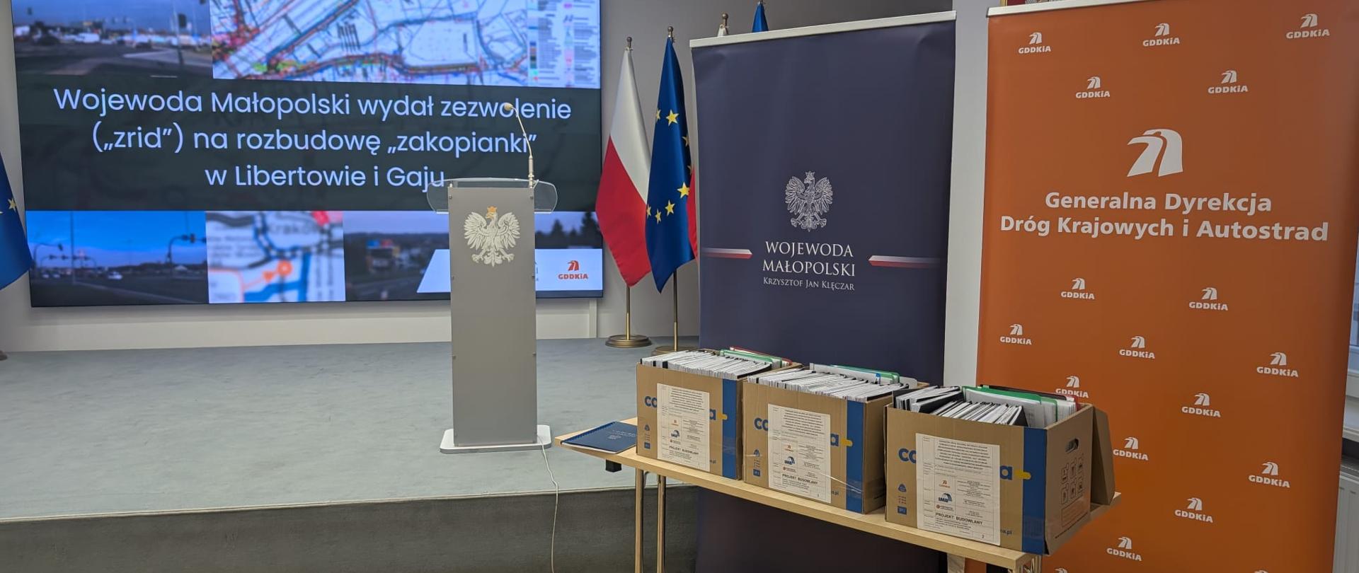 Konferencja prasowa w związku z wydaniem ZRID na rozbudowę DK7 w Libertowie, po prawej roll-upy GDDKiA i Wojewody Małopolskiego, przed nimi dokumentacja w pudłach, na środku zdjęcia podest przygotowany na konferencję