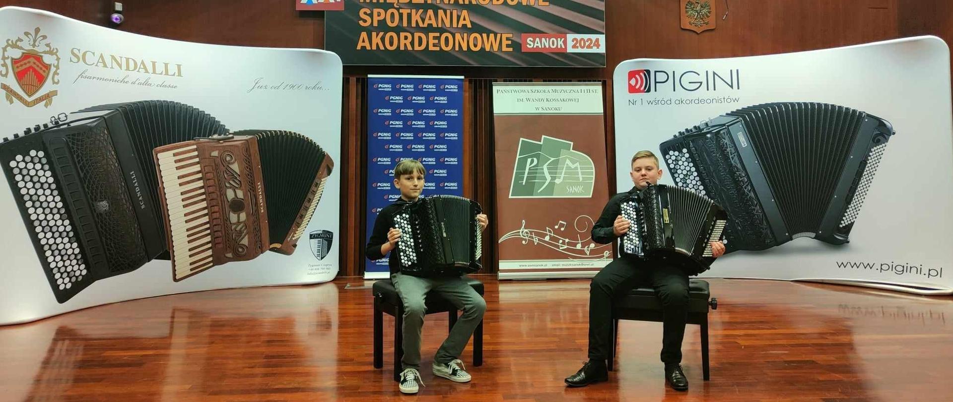 Zdjęcie duetu akordeonowego w czasie prezentacji konkursowej. Uczniowie siedzą z instrumentami na krzesłach na scenie - w tle banery dwóch wiodących marek produkujących akordeony