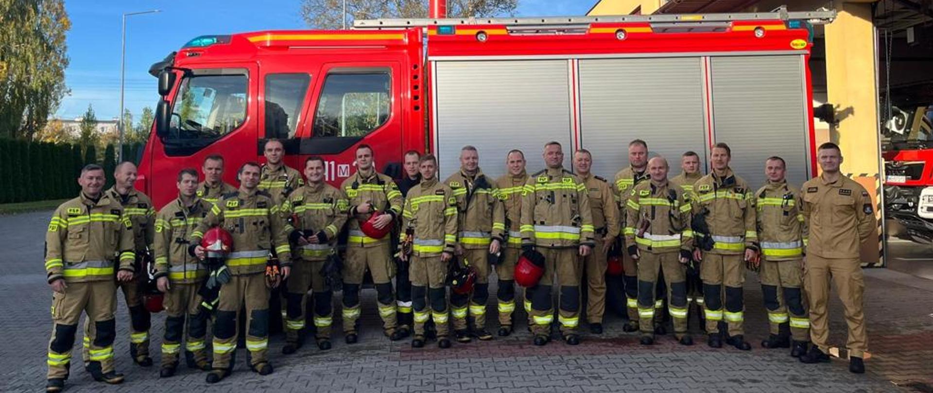 Wspólne zdjęcie strażaków żegnających st. ogn. Marcina Lechowskiego przechodzącego na zaopatrzenie emerytalne na tle samochodu pożarniczego. Strażacy stoją ubrani w ubrania specjalne, 