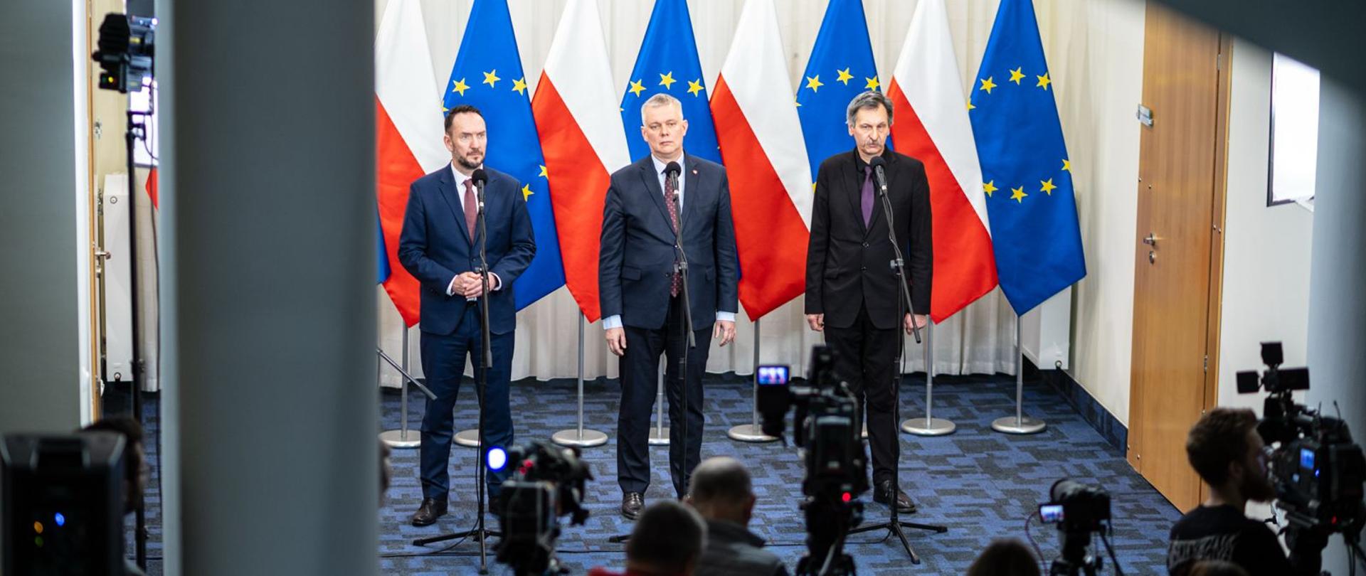 Minister Tomasz Siemoniak, wiceminister Tomasz Szymański oraz dr Grzegorz Kuprianowicz, stoją przy mikrofonach na tle flag polskich i unijnych. Na pierwszym planie kamery i dziennikarze obecni na konferencji 