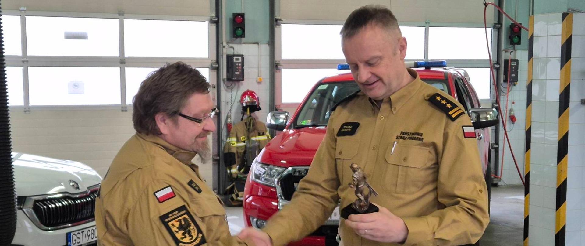 Zdjęcie przedstawia moment wręczenia pamiątkowej figurki strażakowi odchodzącemu na zaopatrzenie emerytalne, w tle samochody pożarnicze.