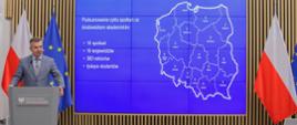 Minister Wieczorek stoi za mównicą i mówi do mikrofonu, za nim na wbudowanym w ścianę wielkim ekranie mapa Polski na niebieskim tle, po bokach flagi Polski i UE.