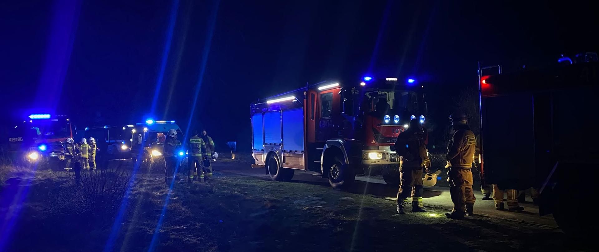 Zdjęcie zrobione nocą. Na zdjęciu samochody strażackie na światłach ostrzegawczych niebieskich, przy samochodach strażacy,