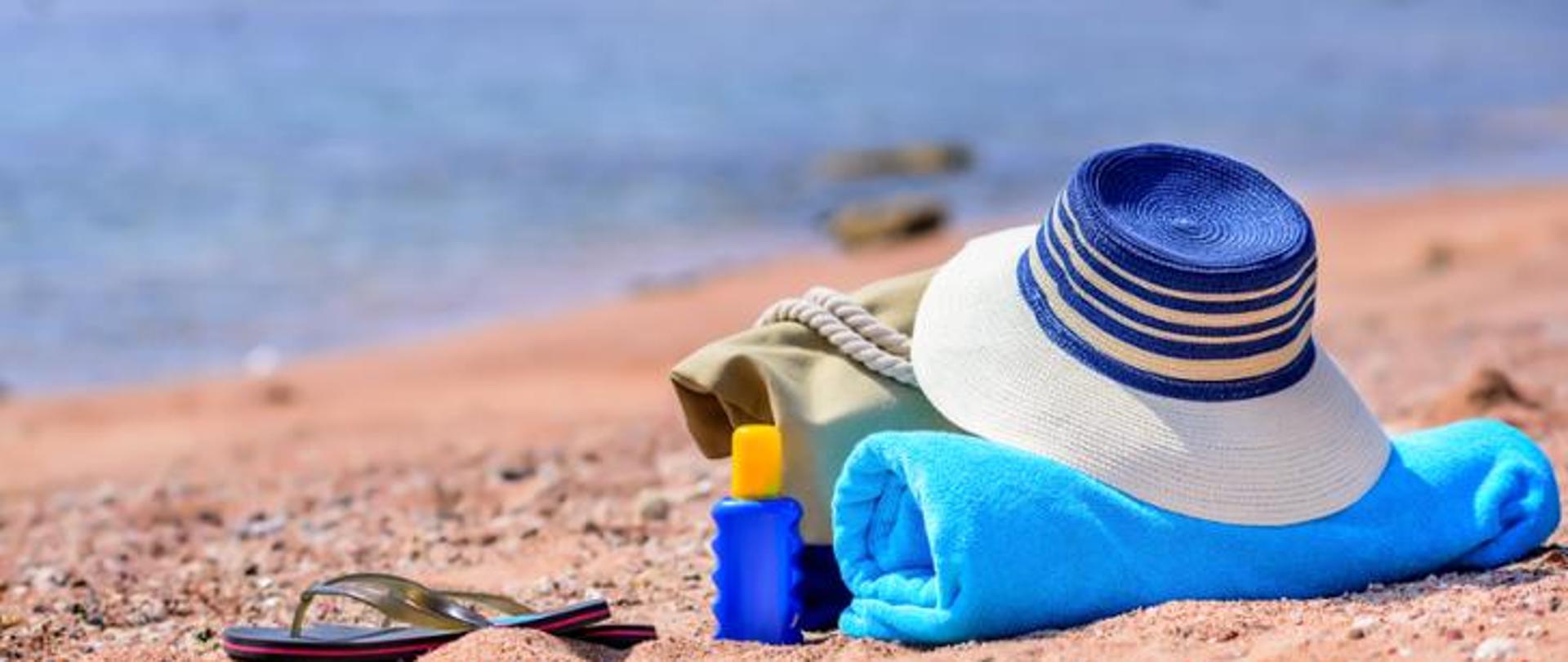 Na zdjęciu znajduje się plaża i morze. Na plaży leży kapelusz, ręcznik, krem przeciwsłoneczny, klapki