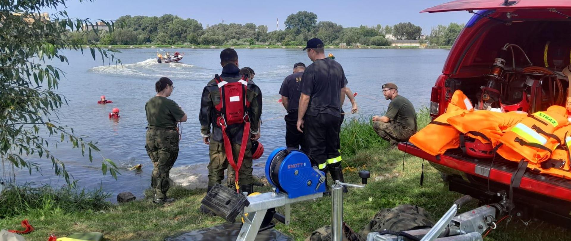 Na zdjęciu akwen wodny na nim jedna łódź strażacka biorąca udział w ćwiczeniach. W wodzie dwie osoby ubrane w ubrania wypornościowe. Na brzegu zbiornika stoją żołnierze i strażacy obserwujący ćwiczenia oraz samochód strażacki z przyczepą do przewozu łodzi.