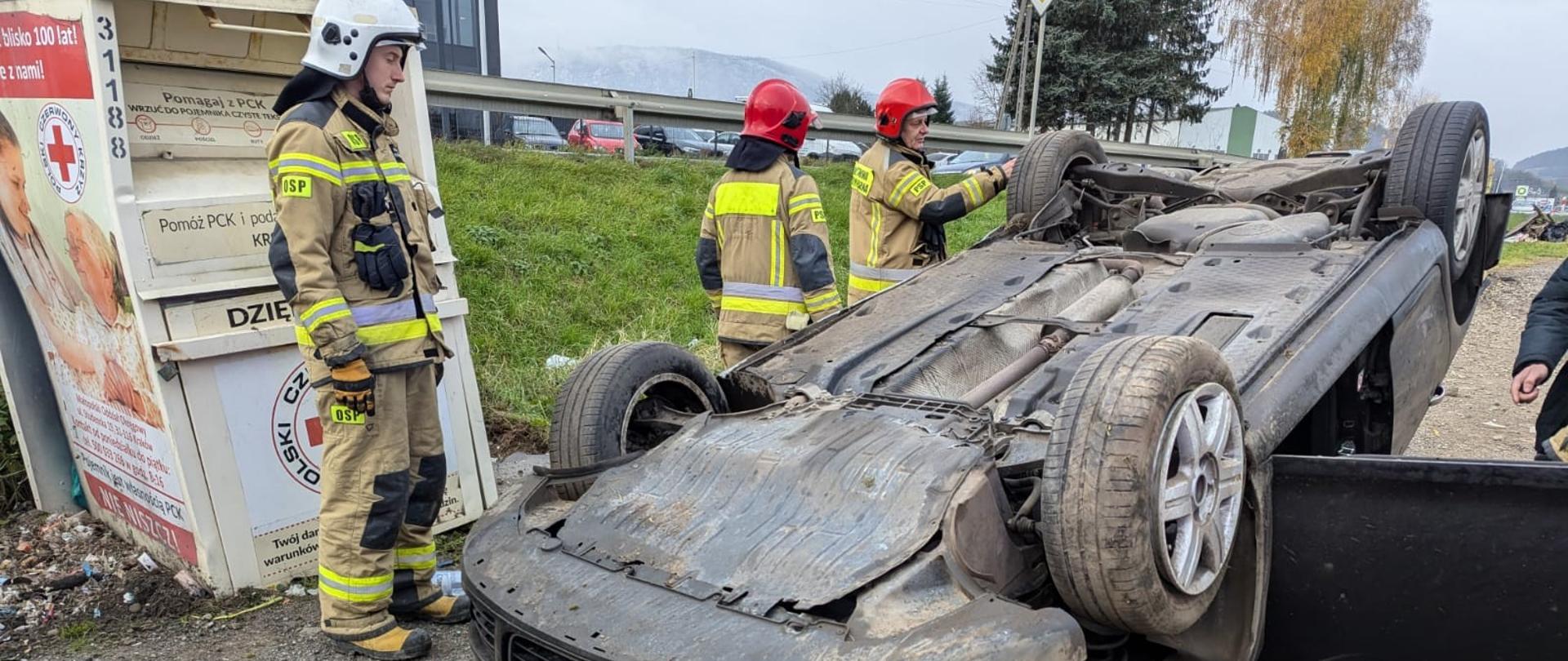 Samochód osobowy na dachu a obok strażacy