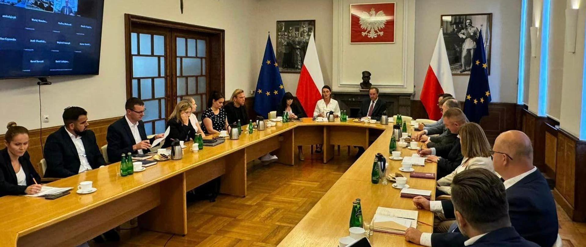 kilkanaście osób siedzi przy okrągłym stole i dyskutuje. w tle f;lagi Polski i UE