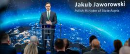 Minister Jakub Jaworowski stoi za mównicą. W tle ekran, na którym widać kulę ziemską. Ujęcie zza słuchaczy.