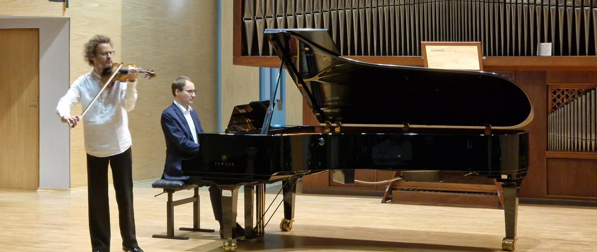 Zdjęcie na scenie sali koncertowej. Skrzypek gra na skrzypcach stojąc, za nim przy fortepianie gra pianista w tle drzwi na zaplecze sali koncertowej. Nad drzwiami krzyż i godło oraz organy.