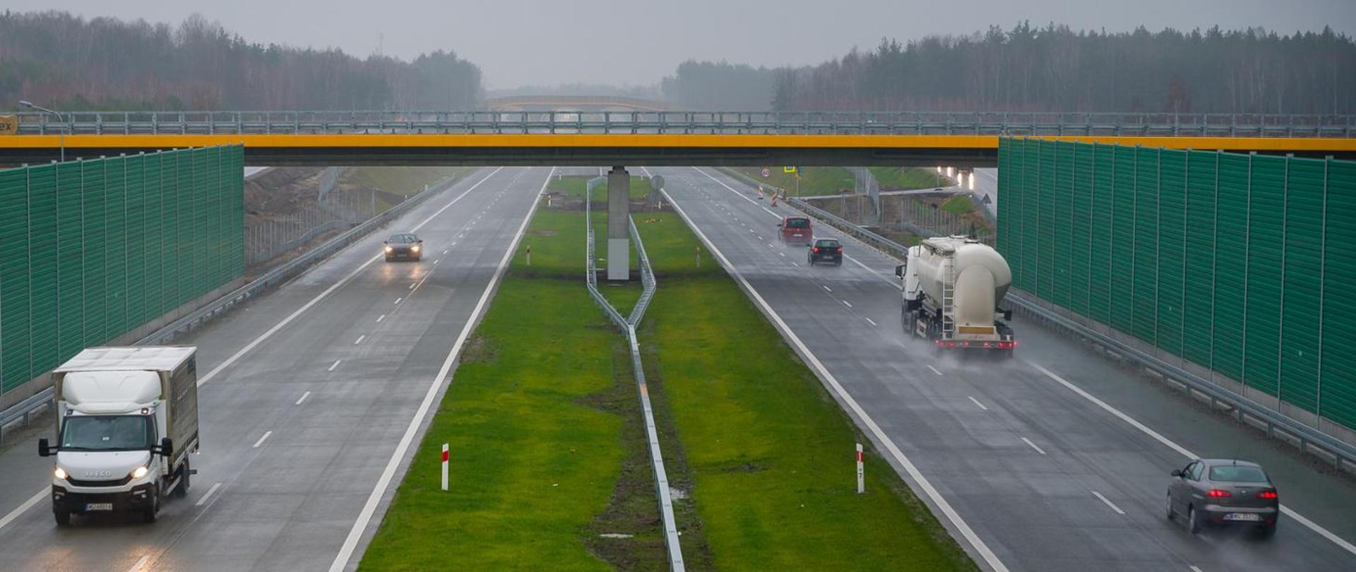 Droga ekspresowa dwujezdniowa, po obu stronach drogi zielone ekrany akustyczne, nad dwujezdniową drogą wiadukt, po obu jezdniach jadą samochody, między jezdniami szeroki pas rozdziału z zieloną trawą. Pogoda deszczowa 