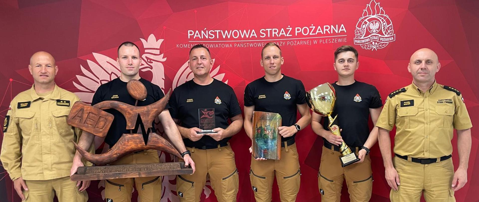 Strażacy z pucharem i statuetką na tle banera komendy powiatowej psp w Pleszewie