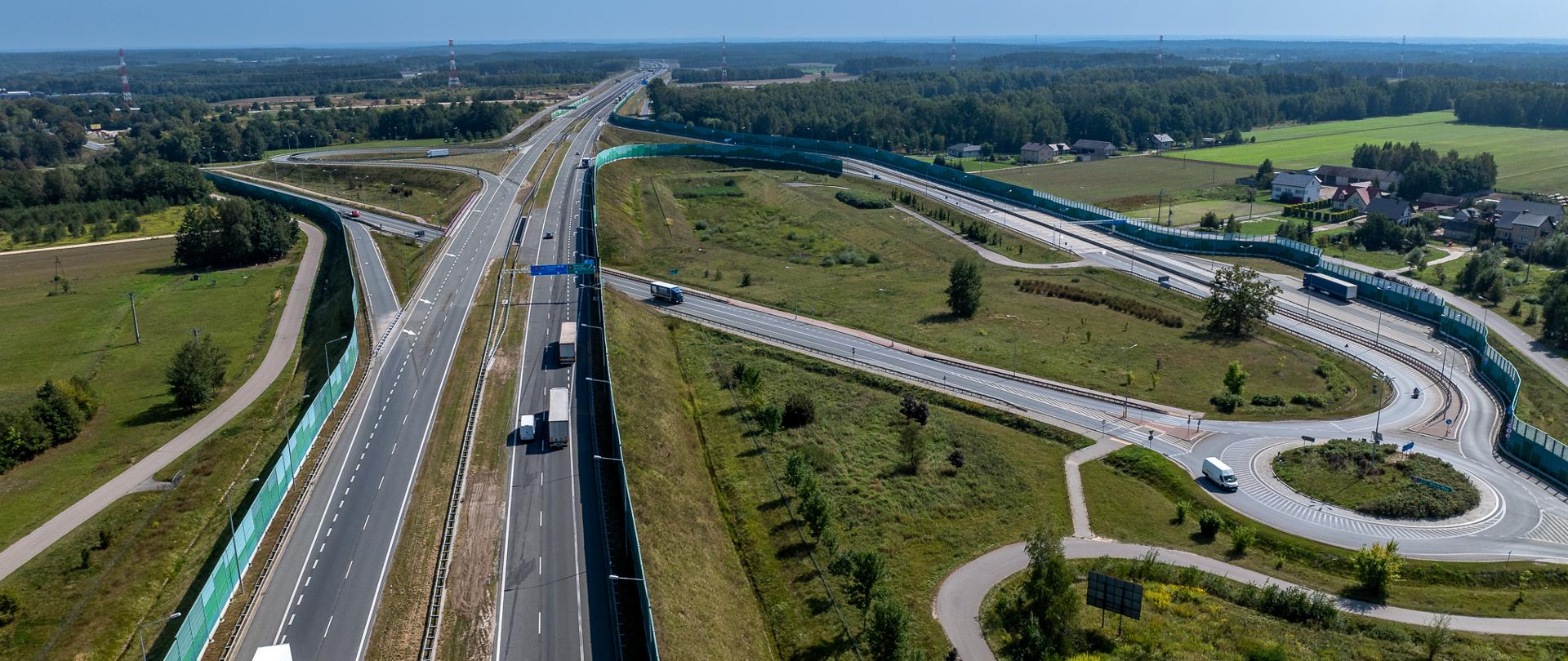 Widok z lotu ptaka na pojazdy poruszające się autostradą A2. Słoneczny dzień, w tle widoczne drzewa i nieliczne zabudowania.