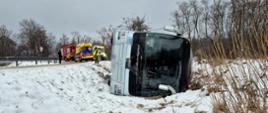 Na zdjęciu widać leżący na boku poza drogą autobus oraz w tle dwie karetki pogotowia i samochód gaśniczy a także funkcjonariuszy różnych służb.