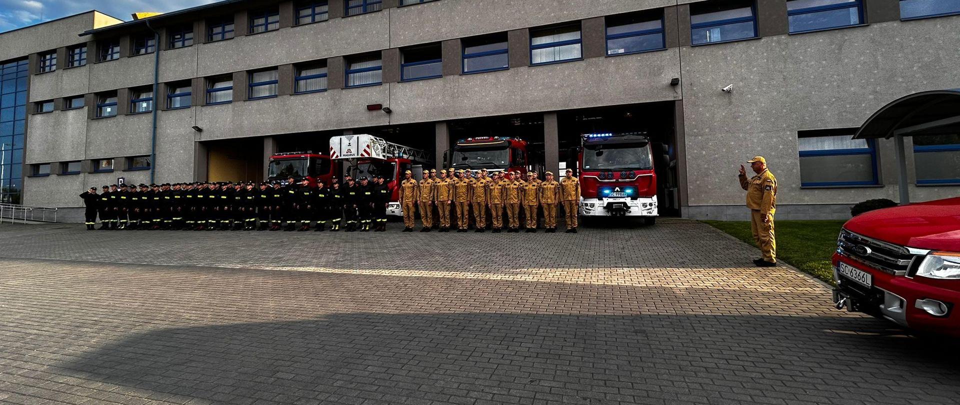 Strażacy stoją w szeregu przed bramami garażowymi jednostki oddając hołd. Za nimi samochody ratowniczo-gaśnicze z włączonymi sygnałami świetlnymi. Po prawej stoi dowódca oddając hołd salutując