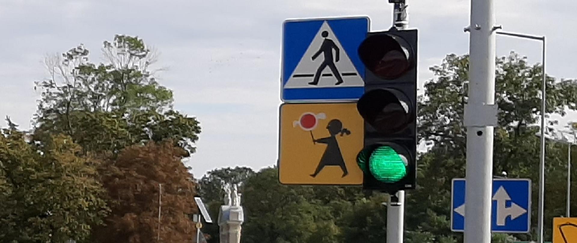 Z prawej strony widoczny sygnalizator z zielonym światłem. Obok na tej samej wysokości znak przedstawiający przejście dla pieszych. w oddali widoczne korony drzew.