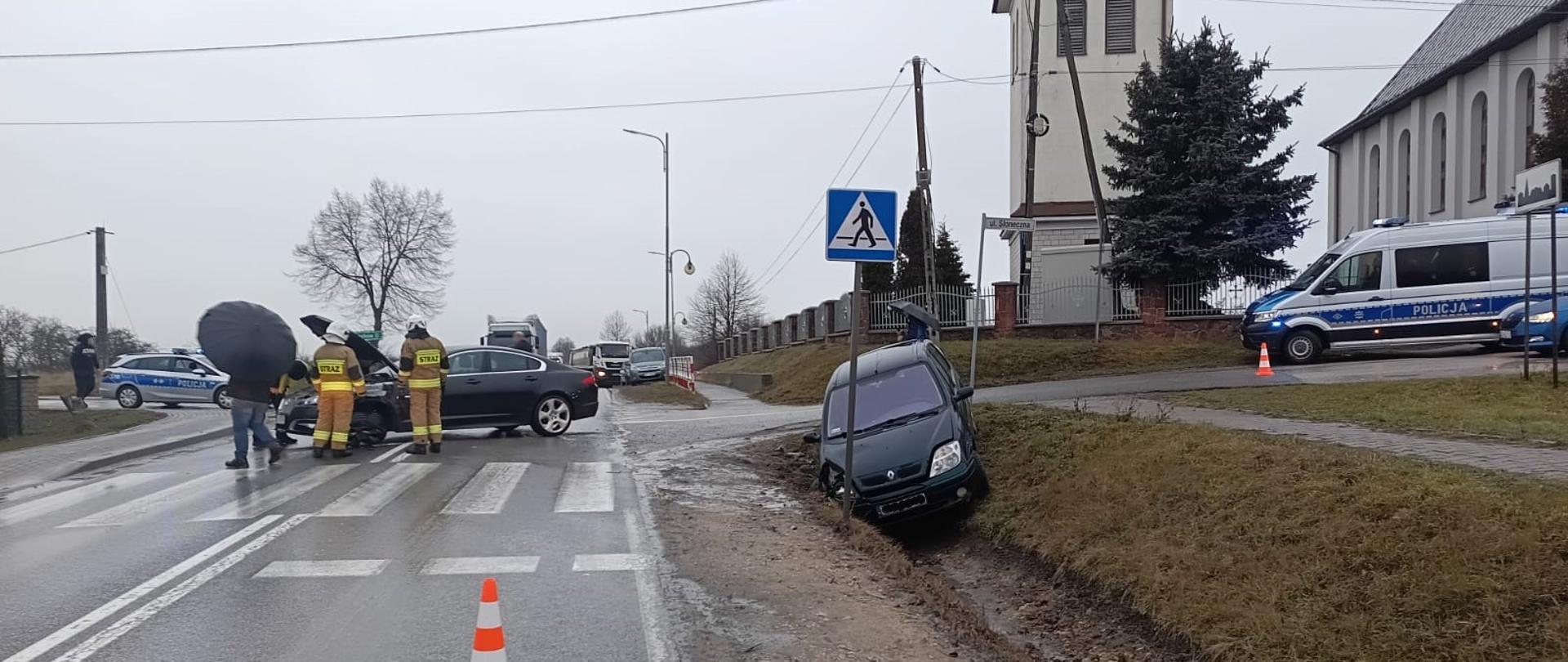 Zdjęcie przedstawia miejsce wypadku: z prawej strony uszkodzony Renault stojący w przydrożnym rowie, a w centralnym punkcie na jezdni stoi Jaguar, przy którym pracują strażacy.