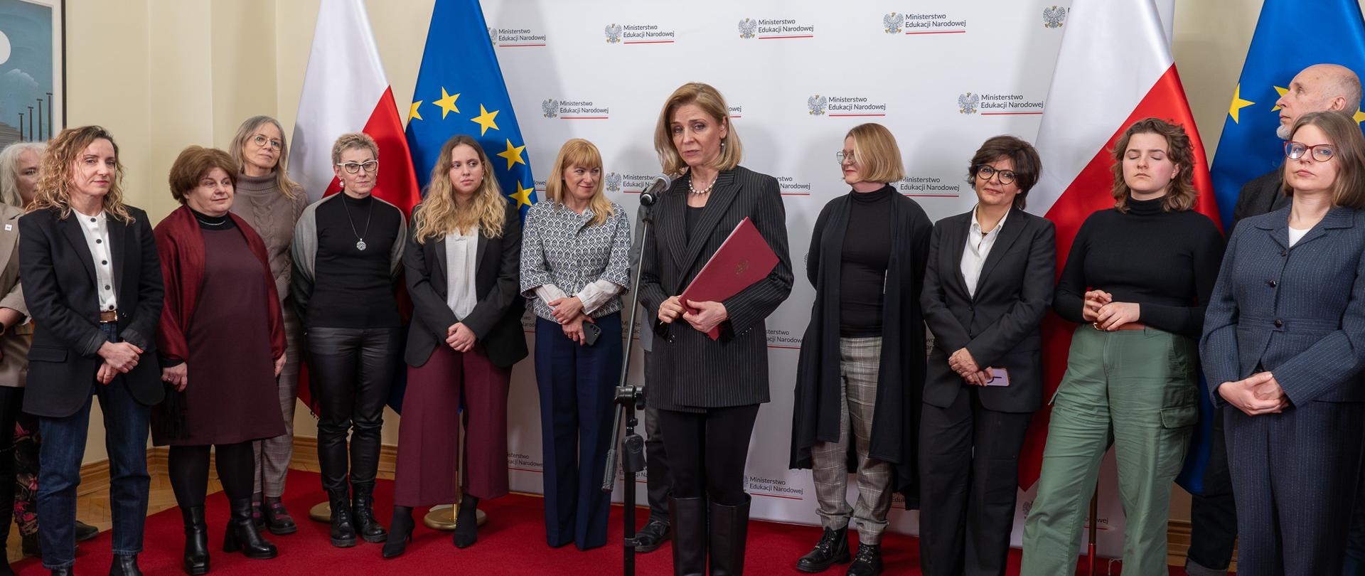 Pod ścianą na tle białej ścianki z napisami Ministerstwo Edukacji Narodowej oraz flag Polski i UE stoi grupa osób, przed nimi przy mikrofonie stoi wiceminister Joanna Mucha 