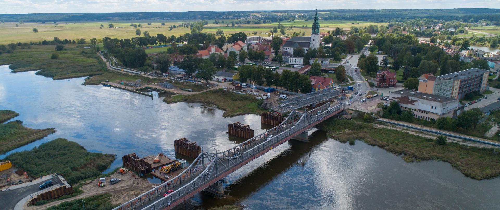 Rzeka, nad nią rozpięty zabytkowy most stalowy. Po drugiej stronie mostu gęsta zabudowa miejska z widoczna wieżą ratuszową. W oddali pola, łąki, a na linie horyzontu ścina lasu.