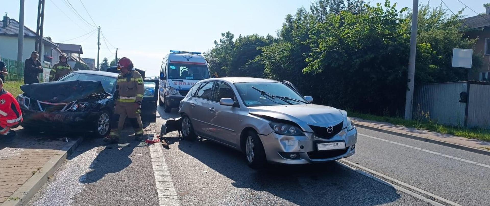 Zdjęcie przedstawia drogę wojewódzką i dwa z trzech aut biorących udział w zderzeniu. Za zniszczonymi autami widać ambulans a między nimi przechodzi strażak.