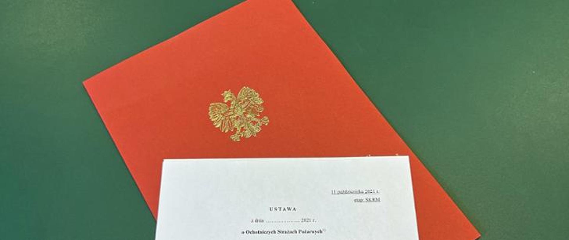 Biała kartka z tytułem dokumentu ułożona na czerwonej teczce papierowej z orłem, pod spodem zielone tło.