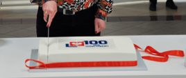 Na stole widoczny biały, prostokątny tort jubileuszowy z logo 100-lecia służby cywilnej, krojony przez pracownicę Naczelnej Dyrekcji Archiwów Państwowych