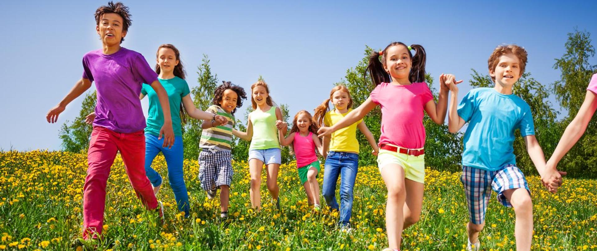 Dziewiątka dzieci biegnie przez zieloną łąkę na której rosną żółte kwiaty. Wszystkie dzieci mają na sobie kolorowe, letnie ubrania i trzymają się za ręce