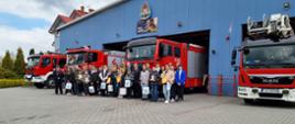 Zdjęcie przedstawia wszystkich laureatów Ogólnopolskiego Turnieju Wiedzy Pożarniczej „Młodzież zapobiega pożarom” wraz z opiekunami. Wszyscy znajdują się przed bramami garażowymi na tle samochodów ratowniczo-gaśniczych. 