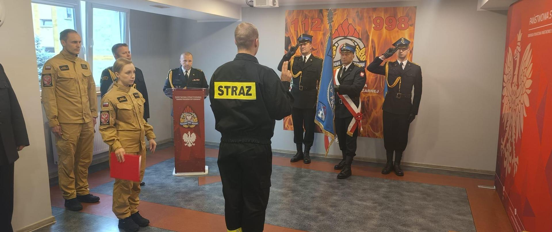 Świetlica strażacka. Trwa ślubowanie strażaka. Na wprost stoi poczet sztandarowy.