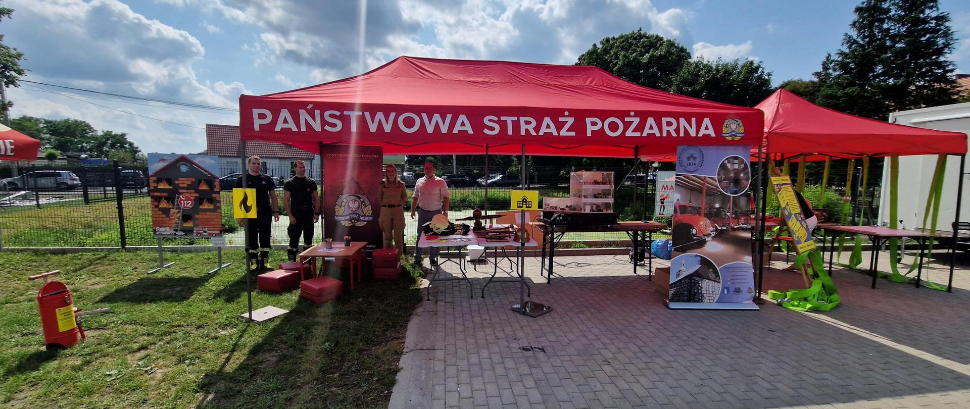 Zdjęcie wykonane na zewnątrz. Ładna pogoda. Na zdjęciu widoczne stoisko Straży pożarnej podczas festynu. Pod namiotem stoją ludzie.