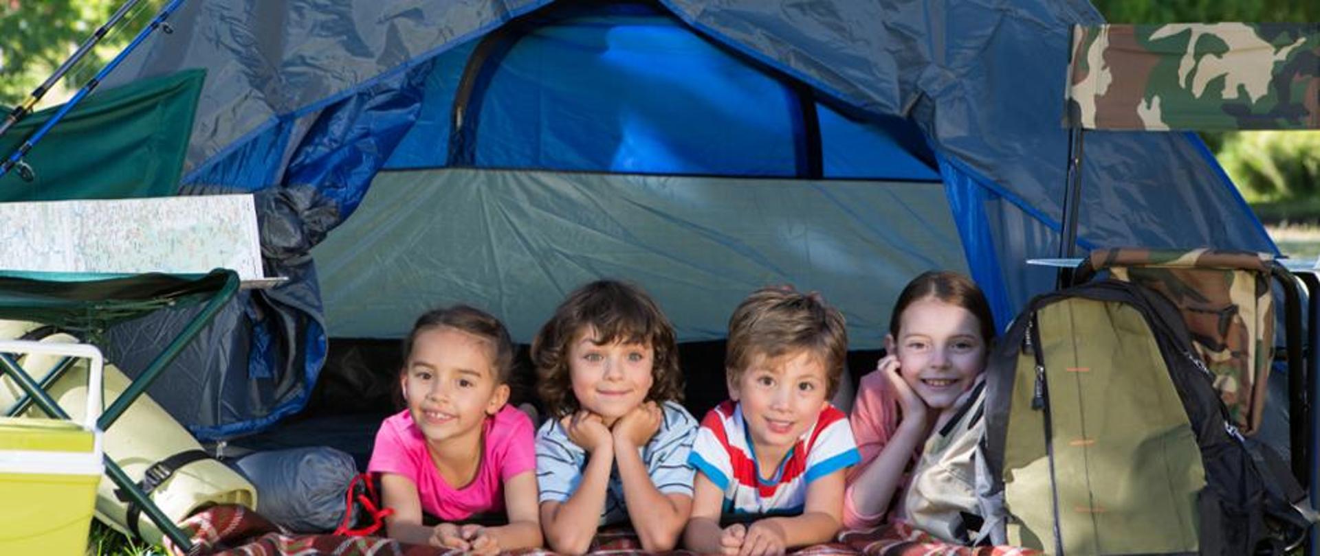 Na zdjęciu znajduje się czwórka uśmiechniętych dzieci leżących w namiocie, po obu bokach namiotu znajdują się akcesoria obozowe: plecak, karimata, śpiwór, składane krzesło 