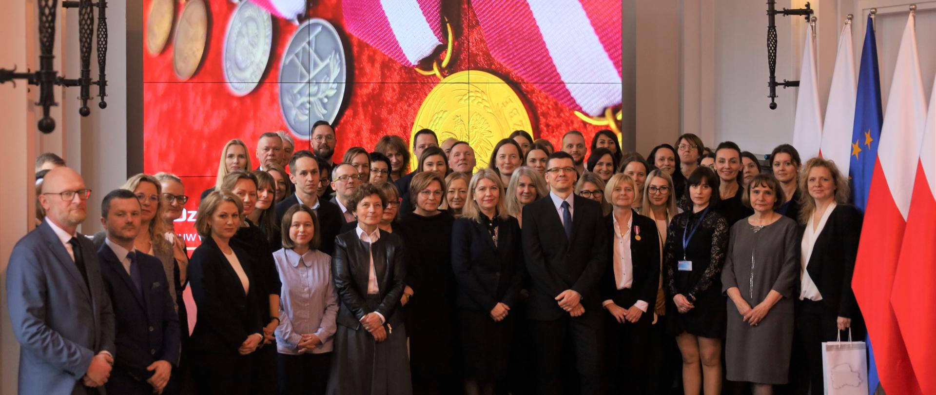 Na zdjęciu widać grupę ludzi - kobiety i mężczyźni. W tle ekran z wyświetlonym zdjęciem medali, po prawej stronie flagi - Polski i Unii Europejskiej 