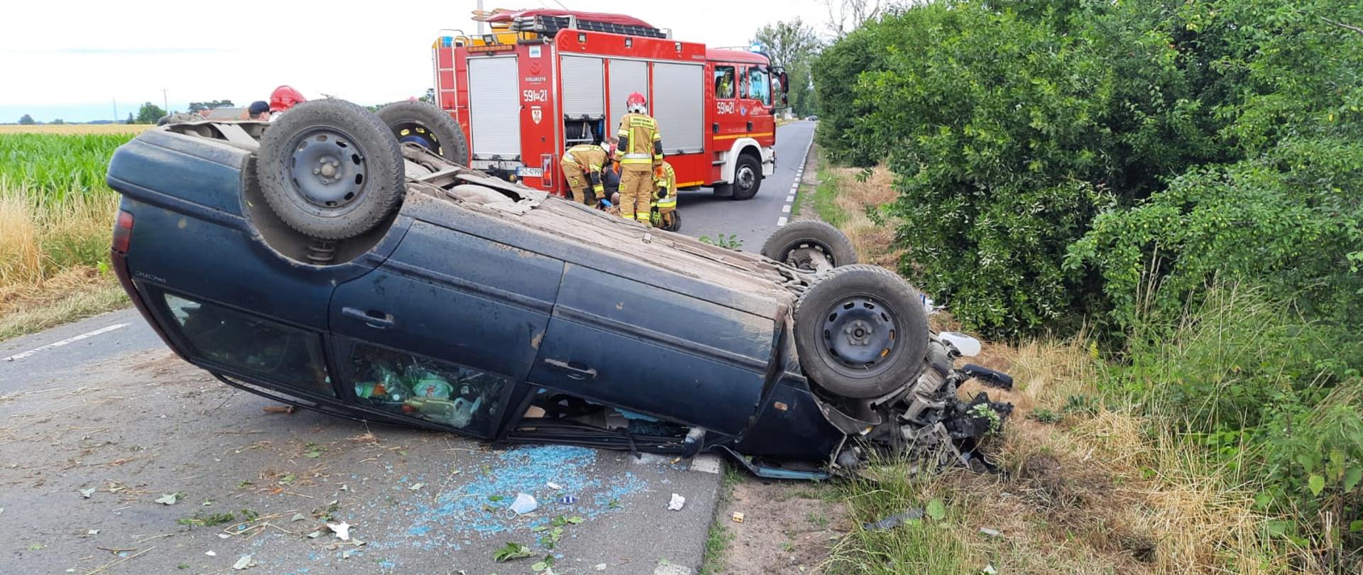 Widać samochód osobowy po dachowaniu na drodze, w tle samochód straży pożarnej i strażacy