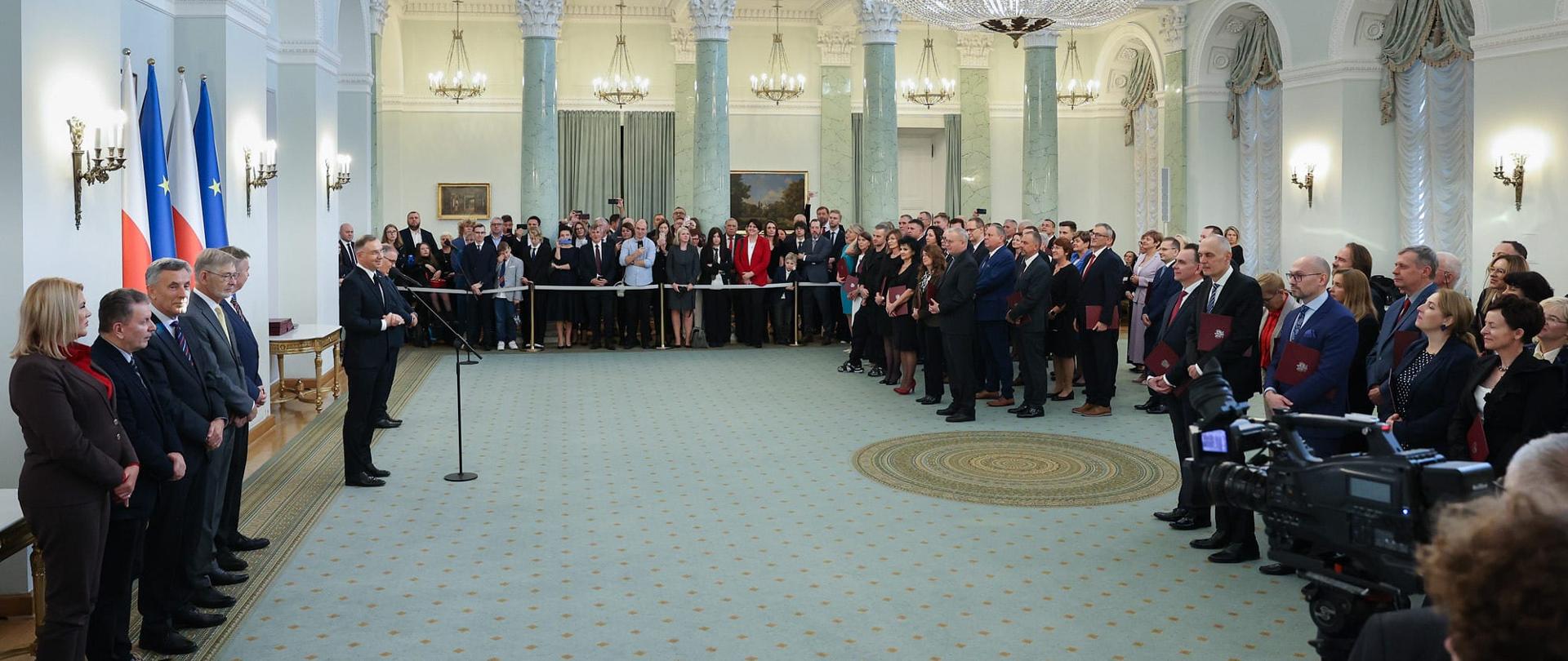 Widok z boku na wielką salę, po lewej stoi prezydent Duda i mówi do mikrofonu na stojaku, naprzeciwko niego po prawej oraz z tyłu sali stoi dużo ludzi, w tle za nimi kolumny.