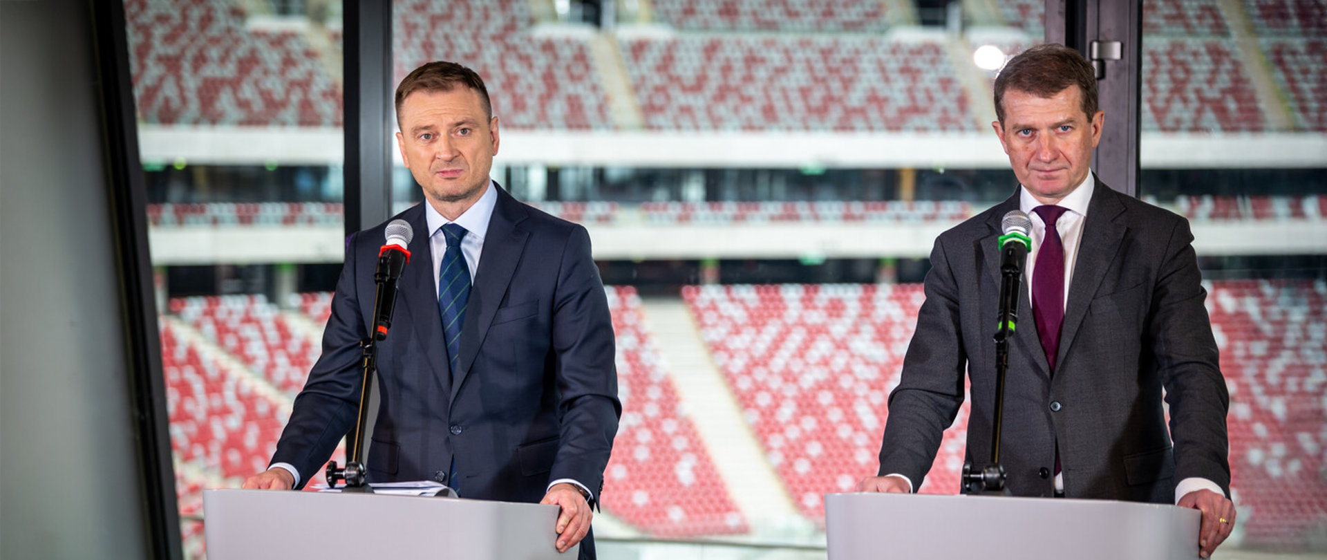 Minister Sportu i Turystyki Sławomir Nitras i Sekretarz Stanu Ireneusz Raś na konferencji prasowej, stoją za mównicami z mikrofonami. W tle za szybą trybuny stadionu PGE Narodowego.