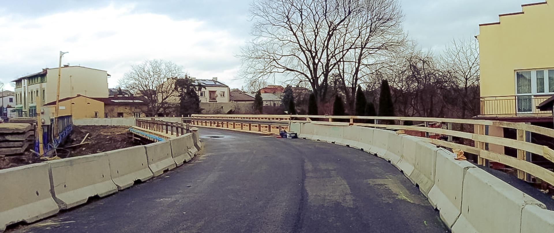 Zdjęcie przedstawia tymczasowy most w trakcie budowy lub remontu. Most jest otoczony betonowymi barierami ochronnymi po obu stronach. Po lewej stronie widać budynki mieszkalne, a po prawej stronie znajduje się żółty budynek z balkonem. W tle widoczne są drzewa bez liści, co sugeruje, że zdjęcie zostało zrobione w okresie jesienno-zimowym. Na drodze znajdują się również narzędzia lub materiały budowlane.