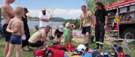 Widok z przodu. Grupa dzieci i młodzieży uczestniczy w pokazach sprzętu ratowniczego. W tle plaża i jezioro. 