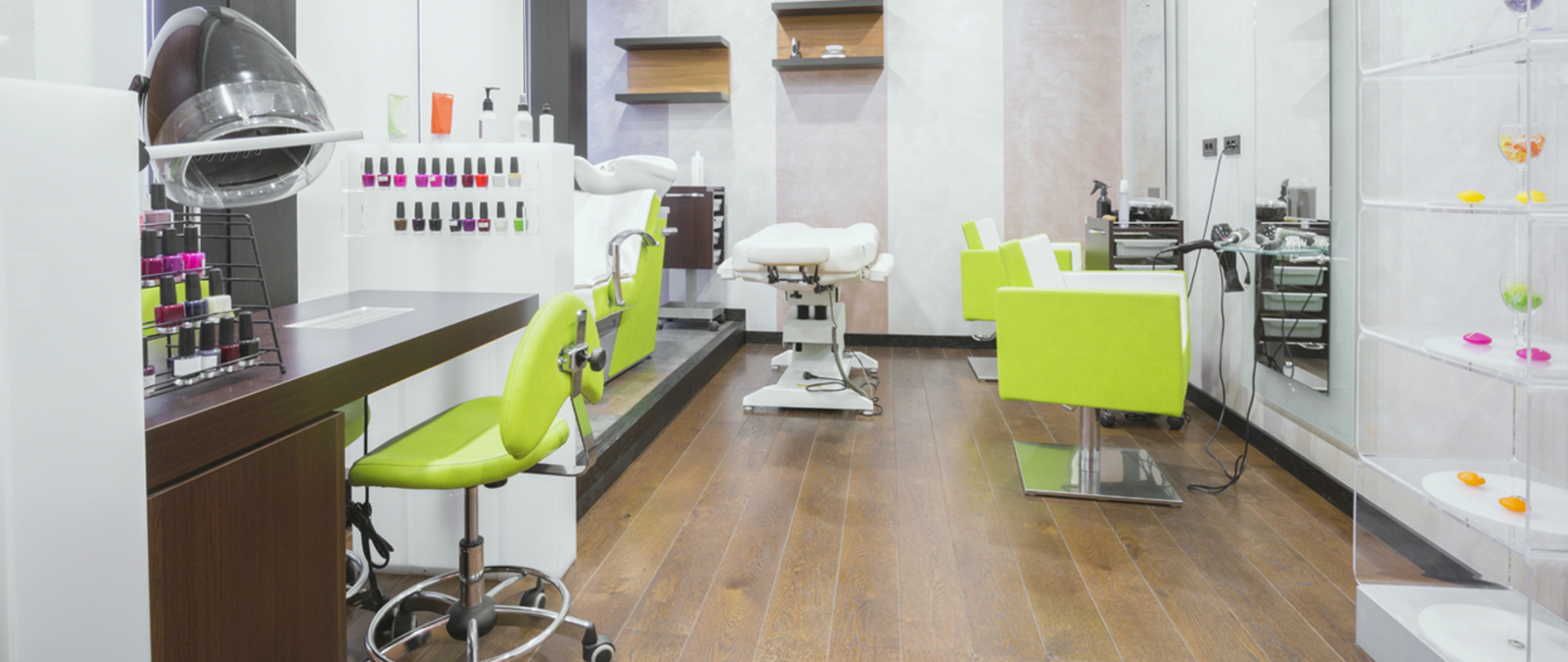 Widoczny oświetlony salon kosmetyczny, w tle fotele dla klientów oraz lampa zabiegowa oraz przezroczysta gablota. Na pierwszym palnie idoczny zestaw lakierów w szerokliej paliecie kolorów wraz lampą. 