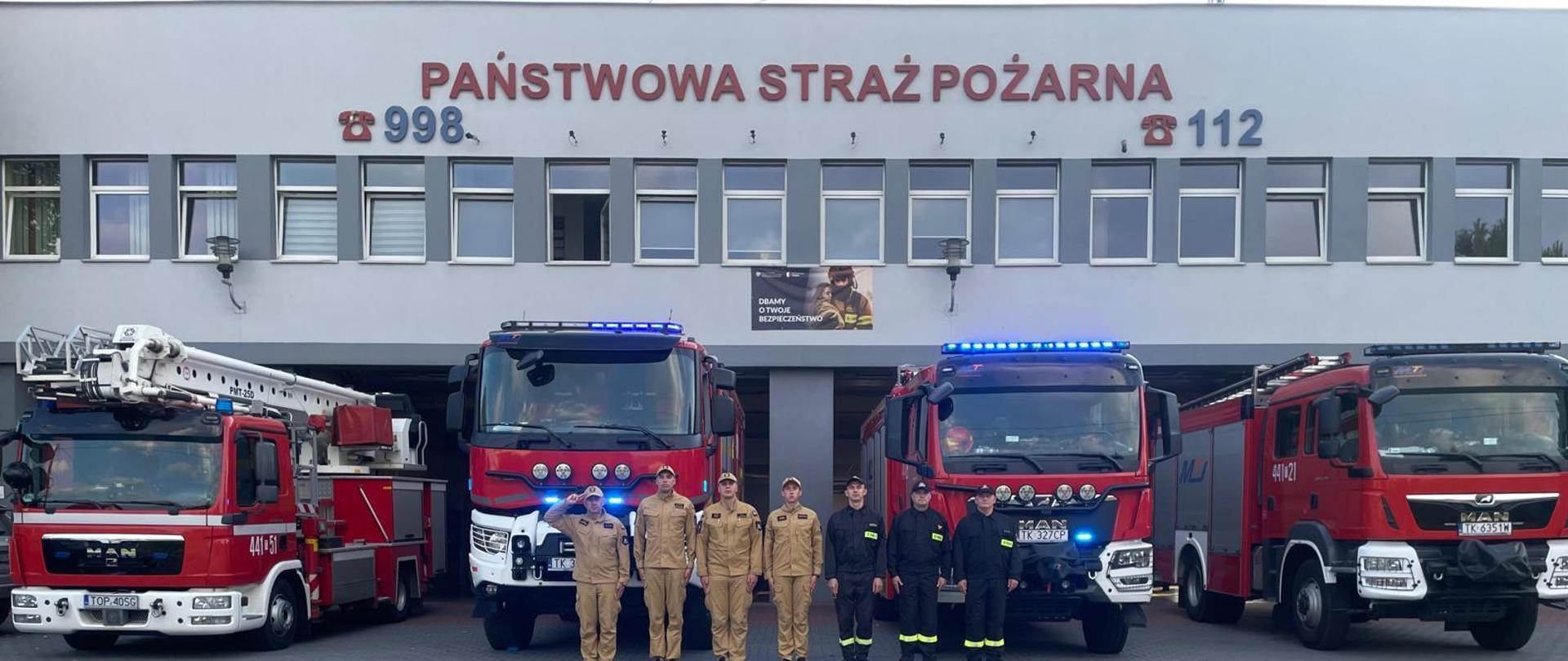 Strażacy stoją w szeregu przed pojazdami pożarniczymi