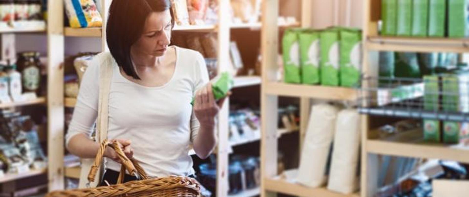 Na zdjęciu widoczna jest kobieta trzymająca koszyk wiklinowy oraz produkt sklepowy, na który zwrócony jest jej wzrok. W tle znajdują się półki sklepowe.