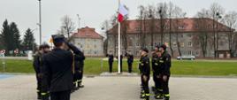 Na zdjęciu znajdują się strażacy podczas uroczystej zmiany służby w czasie której na maszt podnoszona jest flaga państwowa i śpiewany jest hymn państwowy.