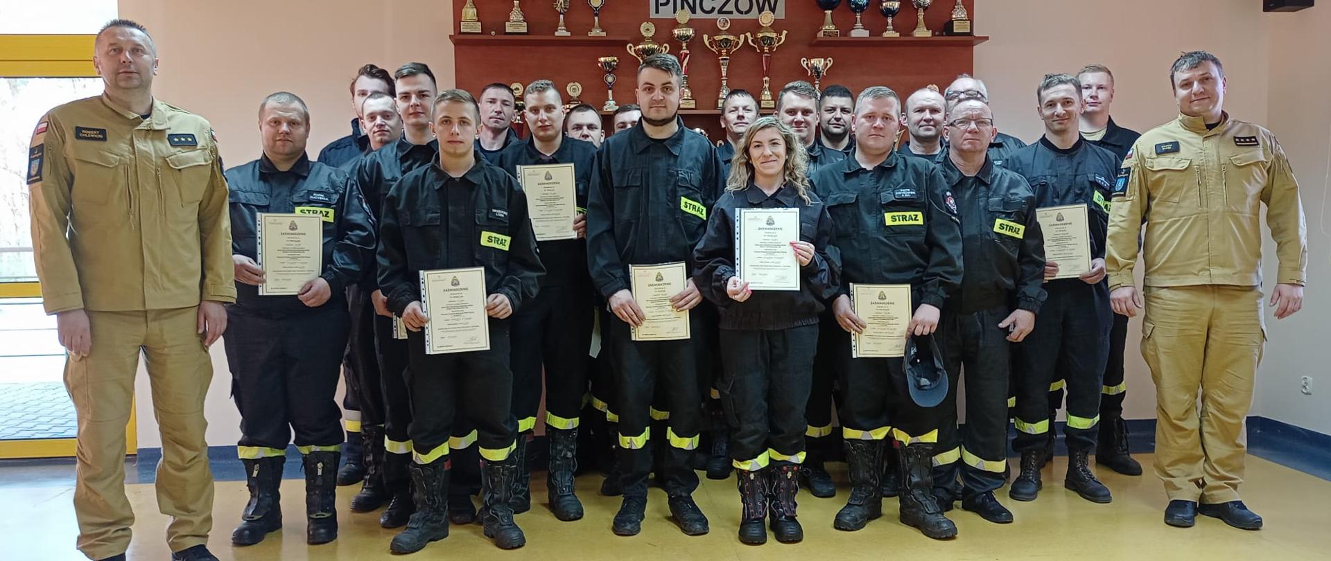 Strażacy - ratownicy Ochotniczych Straży Pożarnych z komisją egzaminacyjną na tle gabloty z pucharami KP PSP w Pińczowie.