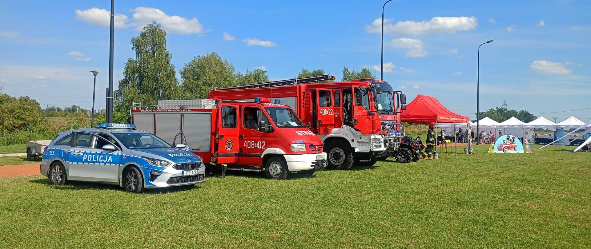 Na trawie stoi radiowóz policyjny oraz 2 samochody pożarnicze, w tle namioty 