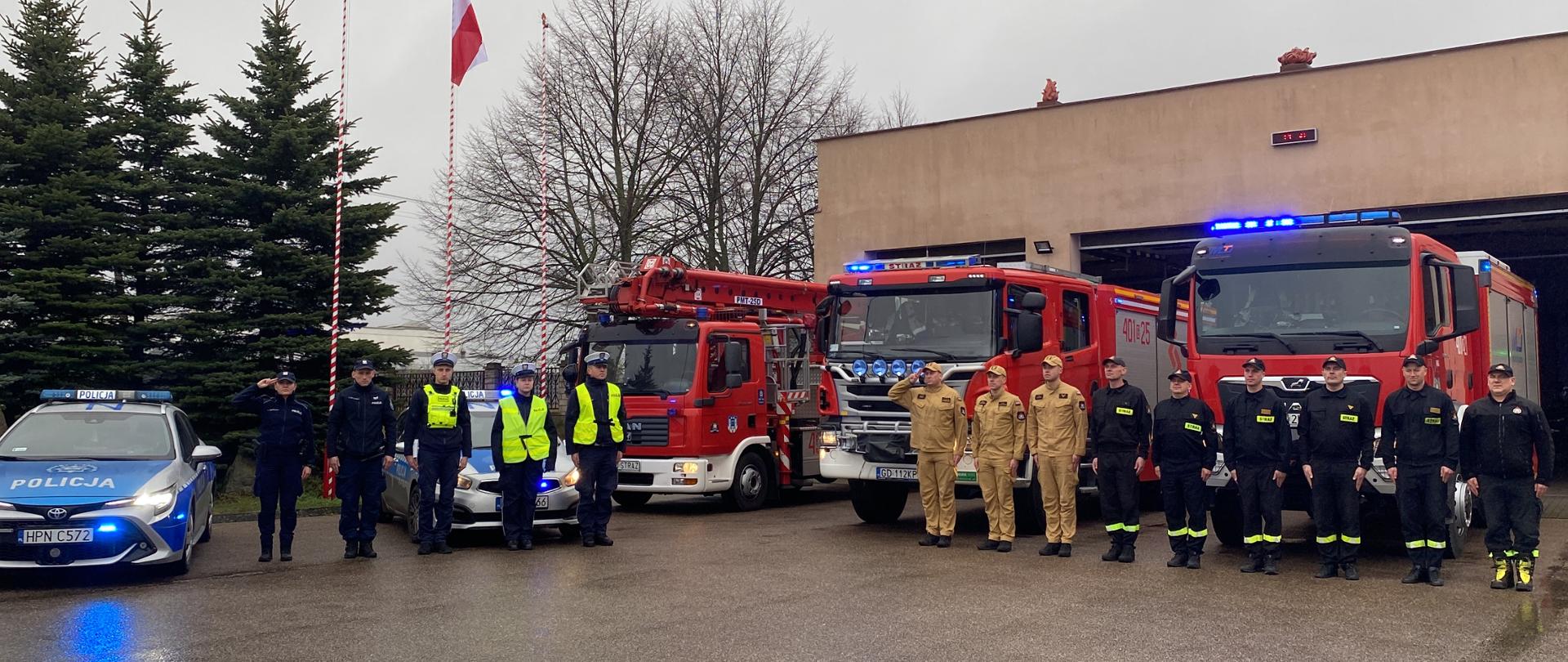 Strażacy i policjanci ustawieni w szeregu na tle pojazdów pożarniczych i radiowozów oddają hołd. 