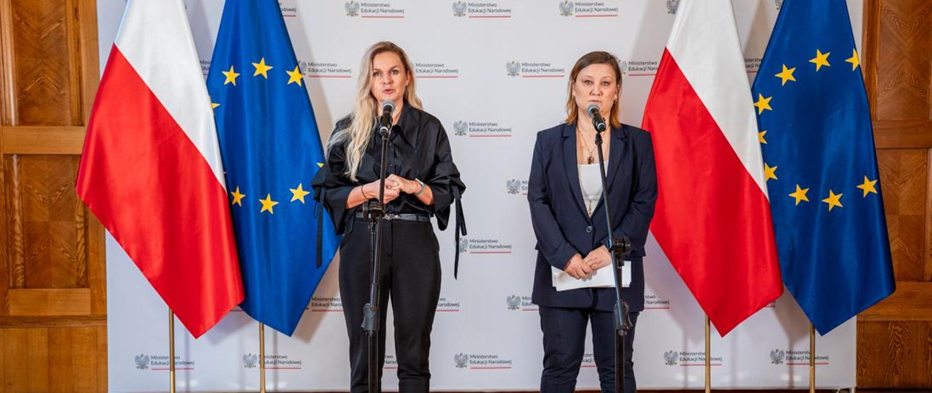 Na tle białej ścianki z napisem Ministerstwo Edukacji Narodowej oraz flag Polski i UE stoi przy mikrofonie minister Barbara Nowacka, a obok niej wiceminister Paulina Piechna-Więckiewicz