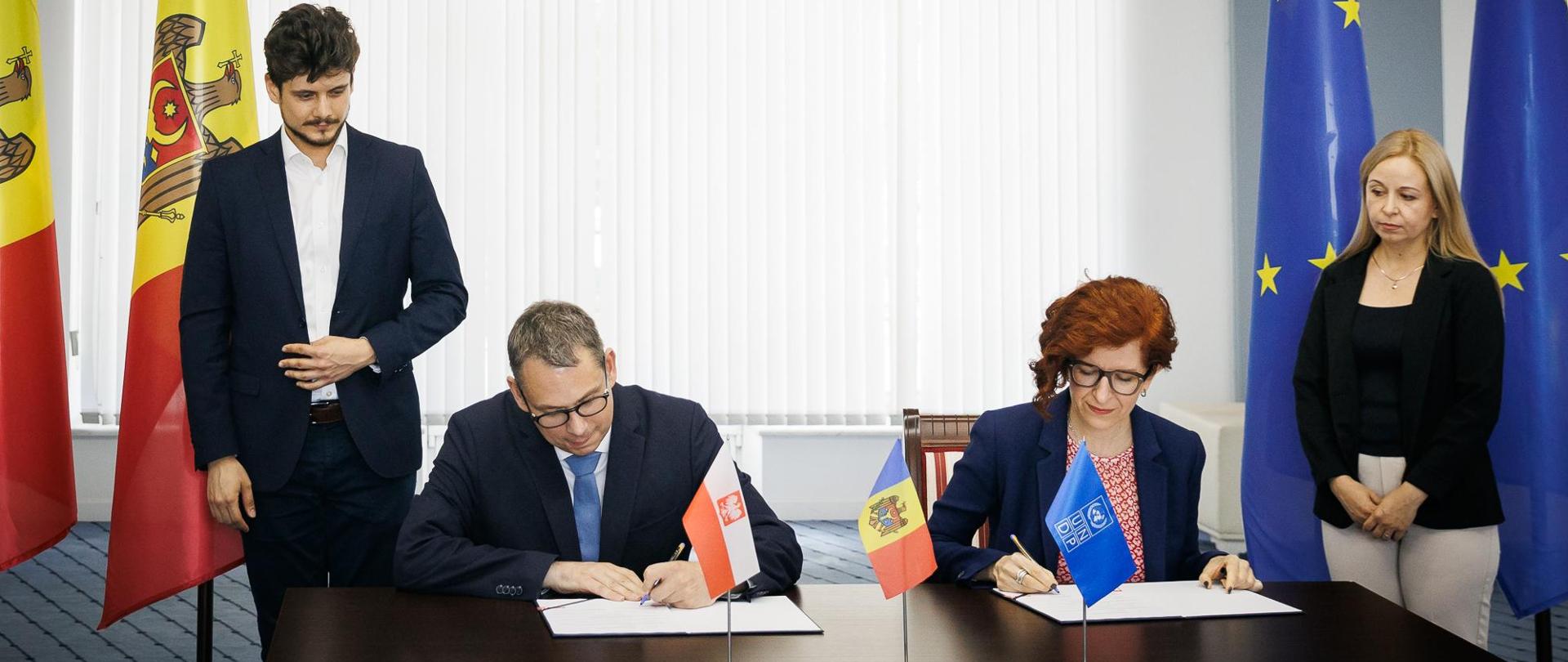 Kobieta i mężczyzna w oficjalnych strojach podpisujące dokumenty przy flagach Polski, Mołdawii, Unii Europejskiej i UNDP.