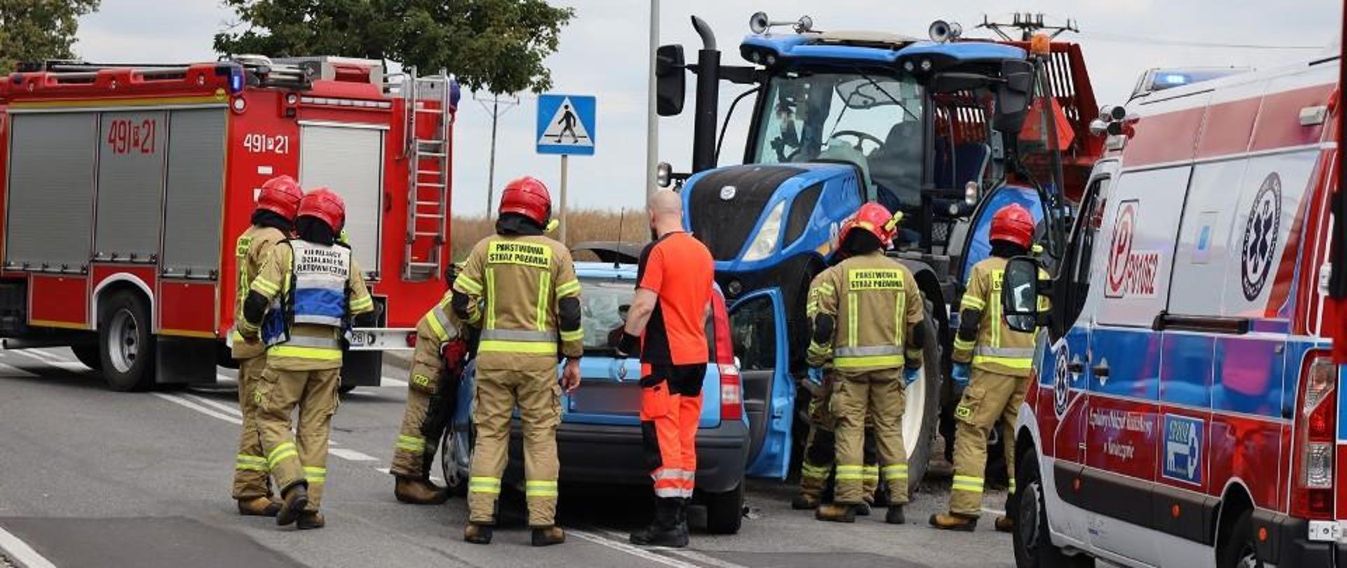 Zderzenie osobówki z ciągnikiem rolniczym w Bożacinie. Strażacy zabezpieczają uszkodzony pojazd osobowy poprzez odłączenie akumulatora. Obok pojazdu stoi również ratownik medyczny, który diagnozował kierowców środków transportu. Droga zablokowana przez pojazdy pożarnicze. 