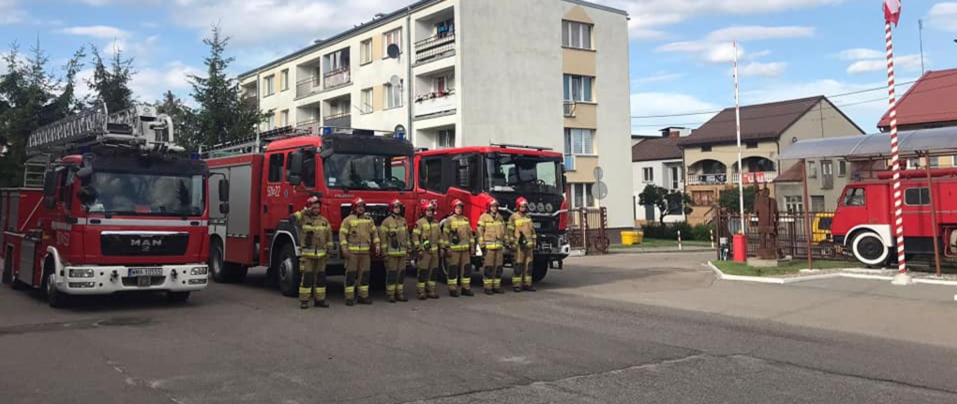 Strażacy stoją przed samochodami strażackimi z włączonym sygnałami świetlnymi i dźwiękowymi na placu komendy.