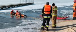 Zdjęcie przedstawia dwóch strażaków asekurujących przy pomocy liny ratowniczej rotę, która działa przy przeręblu wyciągając osobę topiącą się. Na zdjęciu znajdują się również sanie lodowe oraz stojący przy nich strażak