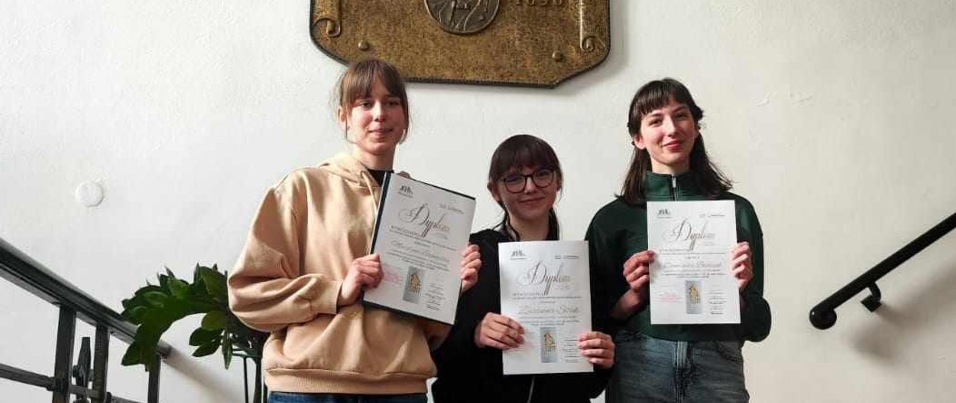 Trzy nastolatki trzymając dyplomy stoją na schodach PSM I i II st, im. Oskara Kolberga.