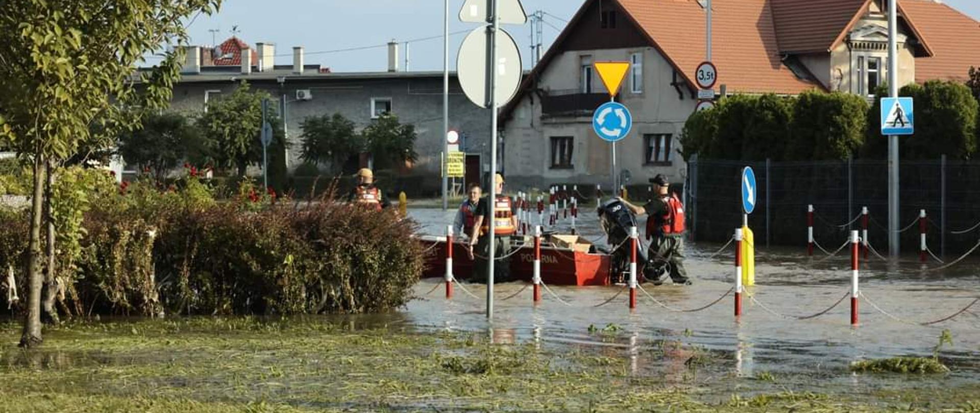 Zdjęcie przedstawia jedną z miejscowości w województwie, które opanowała woda. W tle widać ratowników prowadzących łódź po wodzie. 